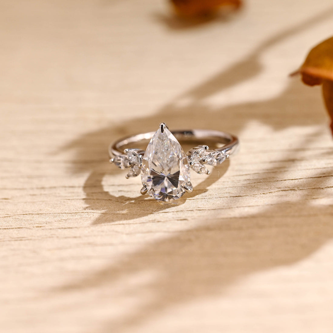 Pear cut moissanite engagement ring with a leaf shaped cluster of marquise stones on each side of the centre stone on a slim polished white gold band
