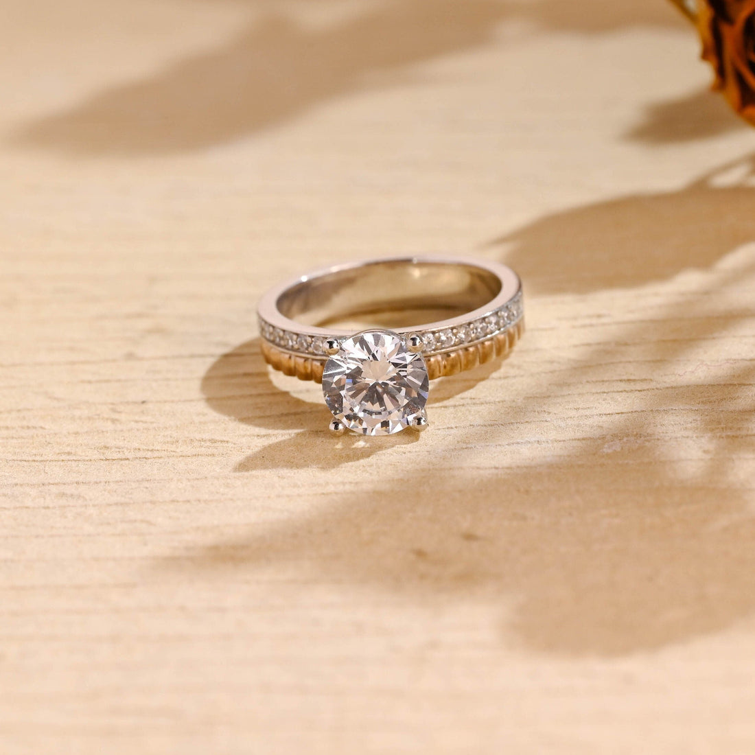 Round moissanite solitaire engagement ring with four claws on a wide band with a row of pave stones and a fluted rose gold edge shown in a two tone finish