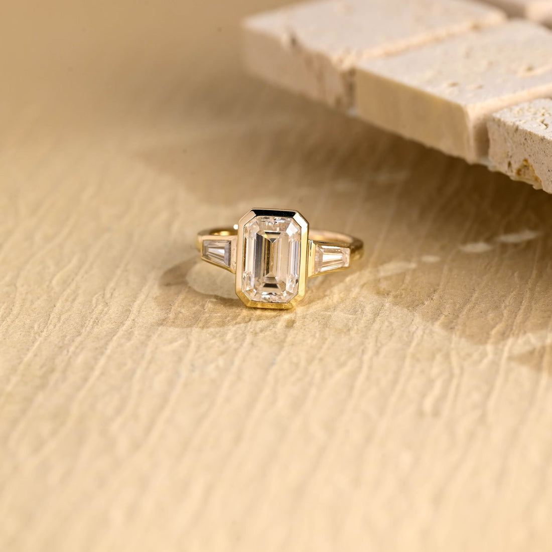 Emerald cut moissanite bezel set three stone engagement ring with a tapered baguette on each side in yellow gold