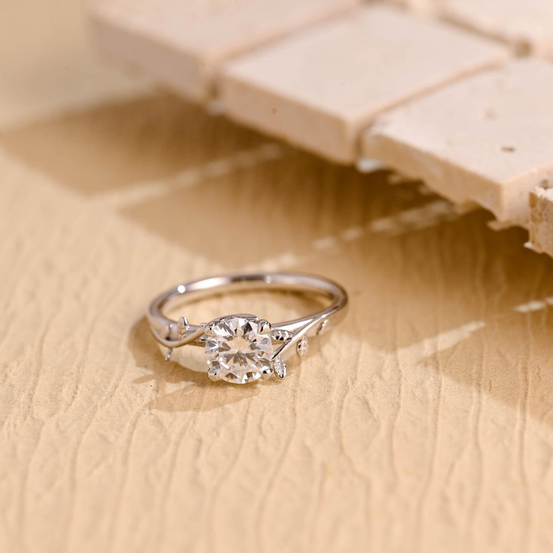 Round moissanite solitaire engagement ring with four claws and a twig shaped white gold band with small leaves on the shoulders