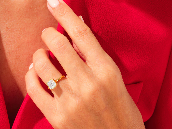 Large Asscher moissanite bezel ring in yellow gold on the ring finger against red fabric
