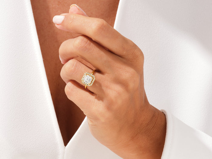 Asscher cut moissanite halo ring in yellow gold worn on the hand against a white jacket