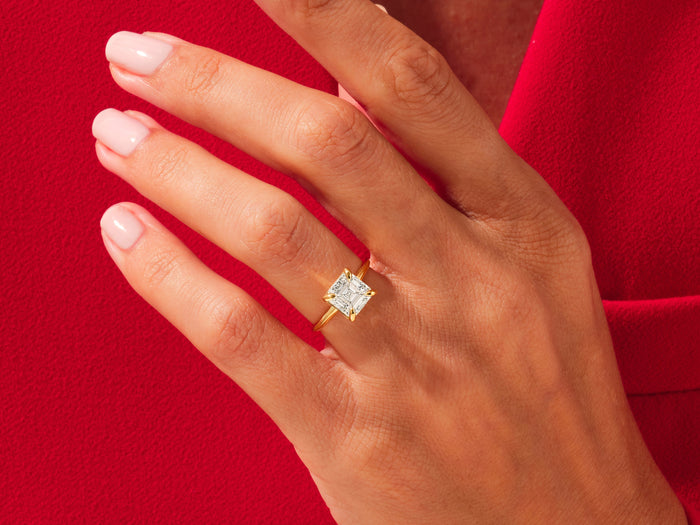 Square asscher lab grown diamond knife edge ring in yellow gold worn on a hand against red fabric