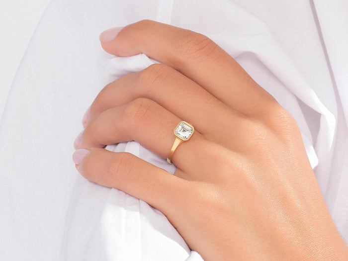 Asscher moissanite bezel engagement ring in yellow gold worn on the hand against a white shirt