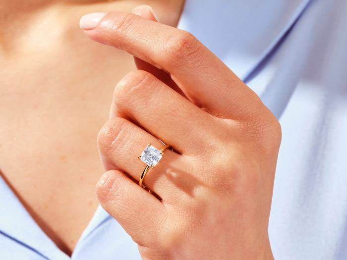 Square Asscher lab grown diamond ring in yellow gold on the ring finger with a blue shirt behind