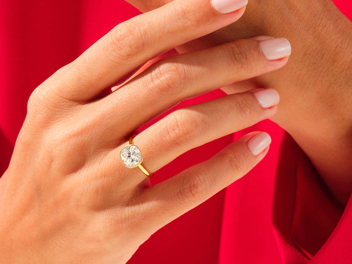 Large cushion moissanite bezel ring in yellow gold worn on the finger against red fabric
