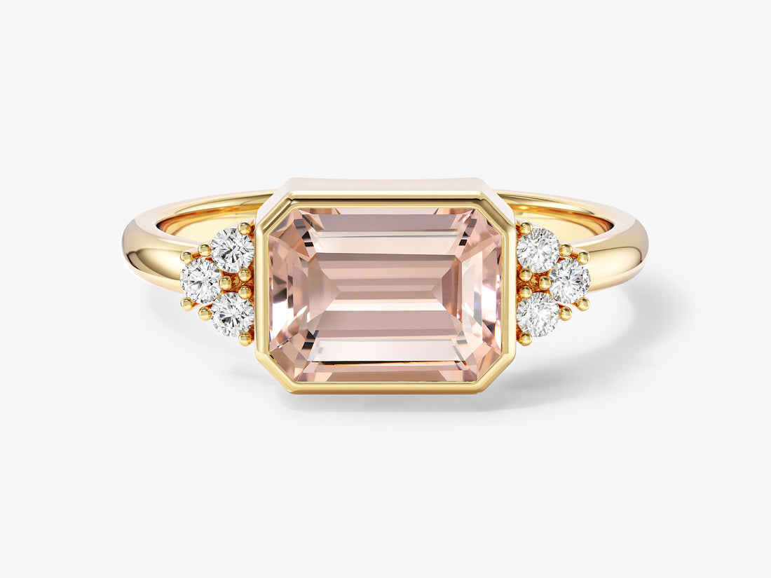 East west emerald cut moissanite bezel engagement ring with round cluster accents in yellow gold, front view, shown with a peach sample center stone