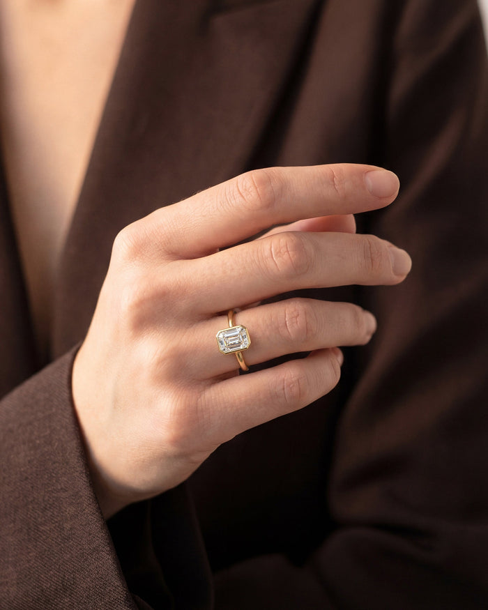 Large emerald cut lab grown diamond bezel ring in yellow gold worn on the hand against a brown jacket