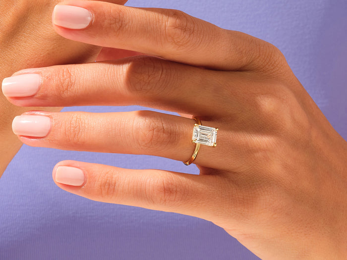 Emerald cut lab grown diamond high set solitaire worn on the hand against a lilac background