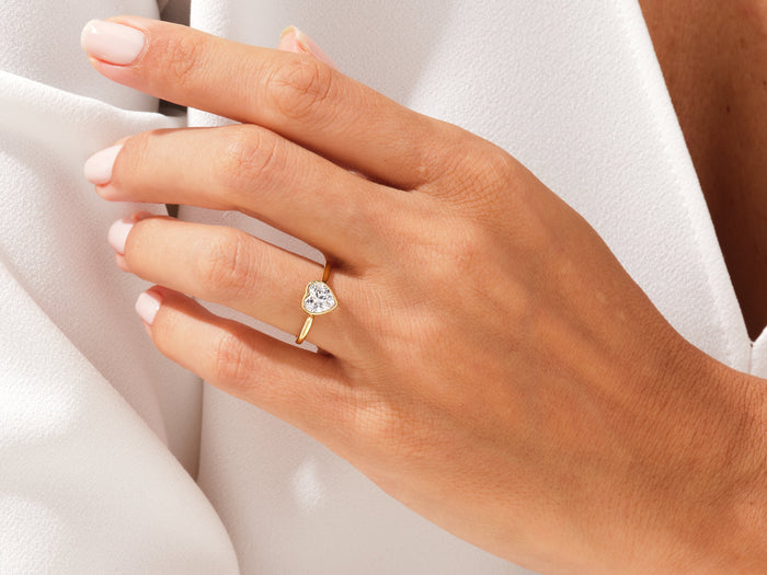 Heart lab grown diamond bezel ring in yellow gold worn on a hand resting on white fabric