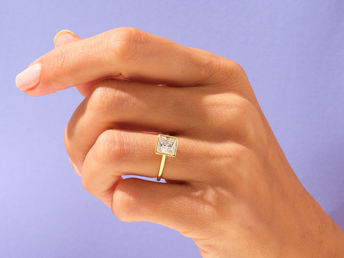 Princess cut lab grown diamond bezel ring in yellow gold on a hand against a lilac background
