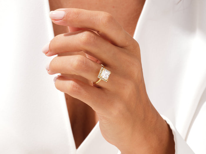 Princess cut lab grown diamond halo ring in yellow gold worn on a hand against a white blouse
