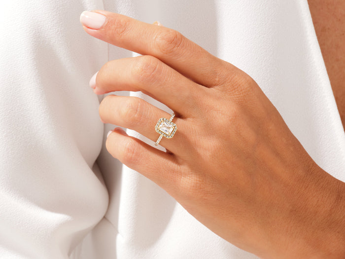 Radiant moissanite halo ring in yellow gold on the ring finger, hand resting on a white shirt