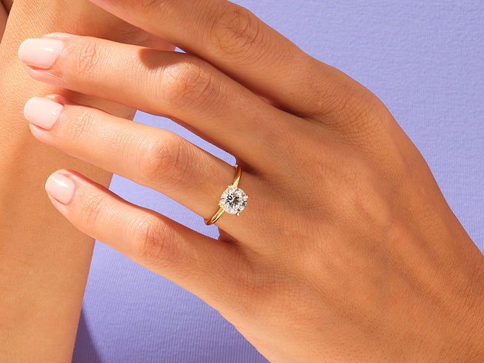Round lab grown diamond knife edge solitaire in yellow gold worn on the hand against a lilac top