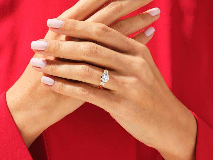 Round lab grown diamond three stone ring in yellow gold on clasped hands against a red top