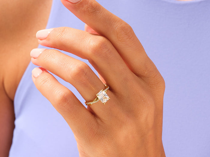 Radiant moissanite rope twist engagement ring in yellow gold worn on the hand against a lilac top