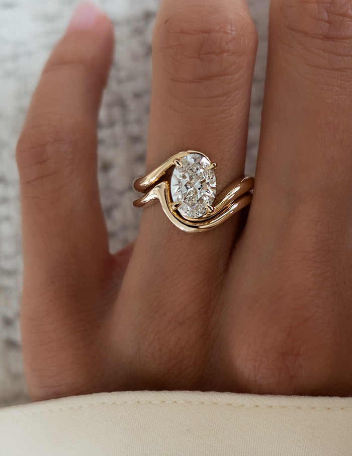 Close-up of oval lab grown diamond swirl ring and plain curved gold band in yellow gold on the hand