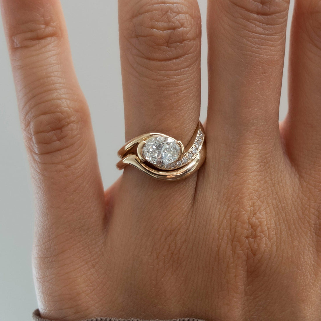 East-west oval moissanite swirl ring with pave arm and plain sculpted wave band in yellow gold on the hand