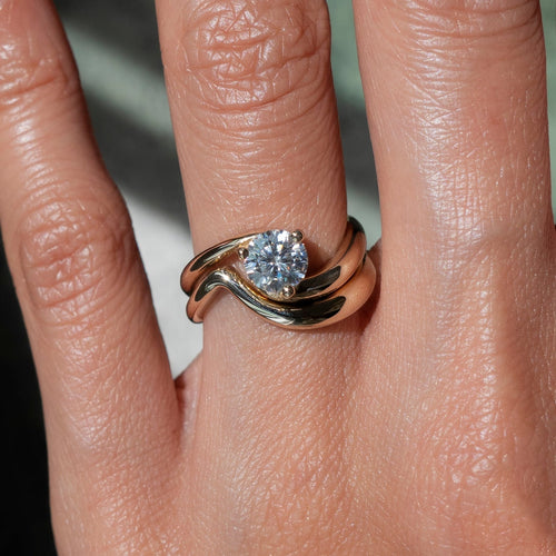 Round moissanite three-prong swirl bridal set with plain wave band in yellow gold on the hand